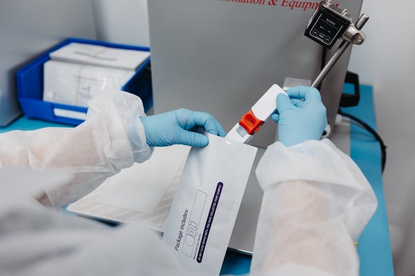 First-person perspective of blue-gloved hands placing an injection-molded diagnostic test kit into packaging.