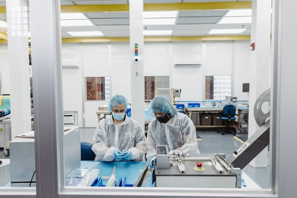 Looking through a window into a Class 8 clean room where two people dressed in coveralls, gloves, face masks, and hairnets work on injection molded plastic test kits for laboratory and at-home diagnostic use.