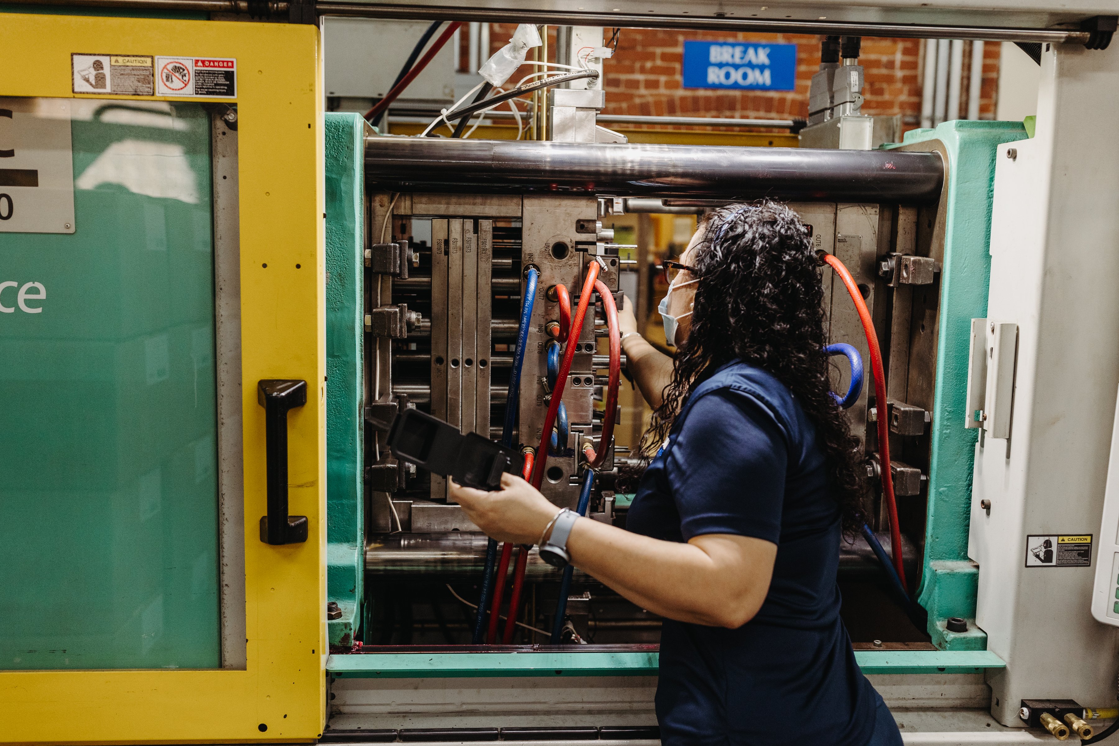 A woman (facing away from the camera) points at the wires in an injection molding machines