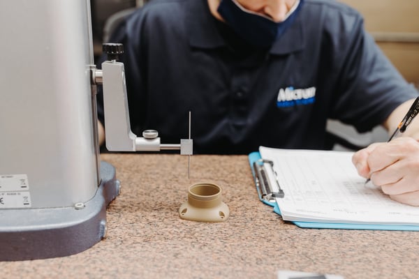 A person in a blue shirt writes on a clipboard during a CMM component inspection. The brown, injection-molded plast