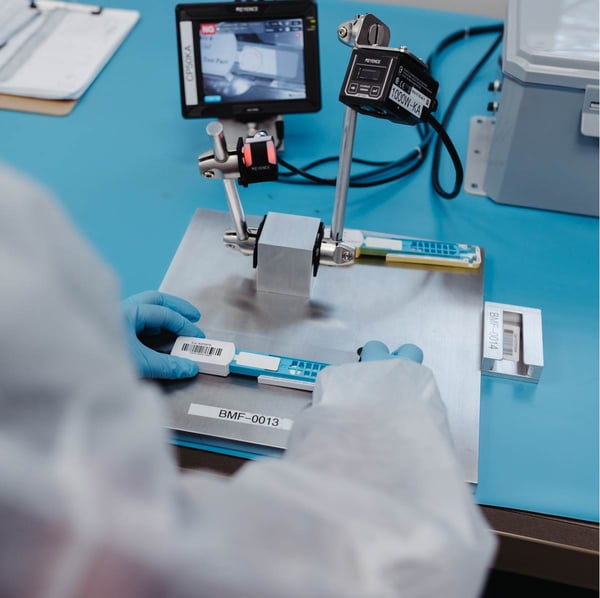 A first person perspective of quality control testing; blue-gloved hands hold an injection-molded plastic part from a diagnostic test kit produced at Micron.