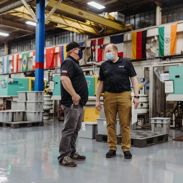 Two employees talk on the manufacturing floor with Arburg injection molding machines in the background